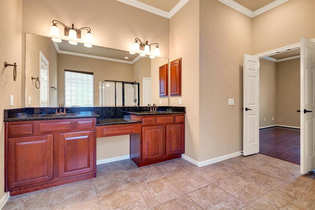 This photo features a spacious bathroom with dual sinks set in a dark granite countertop. Elegant wooden cabinetry provides ample storage, and the room is well-lit with stylish fixtures. The beige walls and tile flooring create a warm, inviting atmosphere. A doorway leads to what appears to be a bedroom with dark wood flooring, enhancing the luxurious feel.