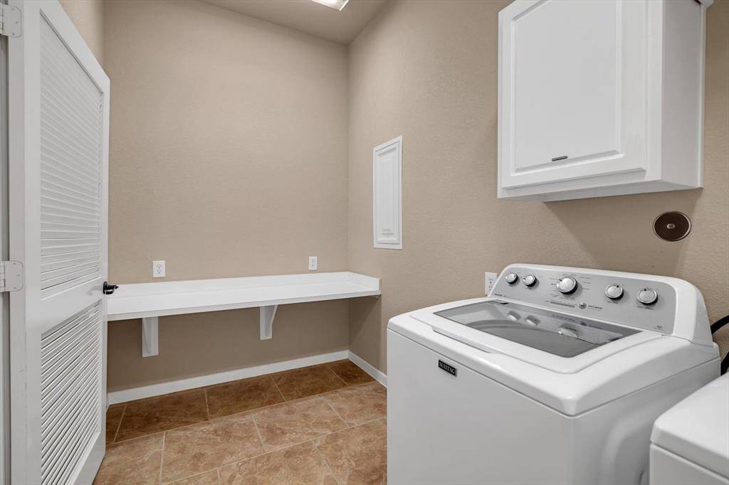 This is a spacious laundry room featuring a modern washing machine, ample counter space, and a wall-mounted cabinet for storage. The room has neutral-colored walls and tiled flooring, providing a clean and functional space for laundry tasks.