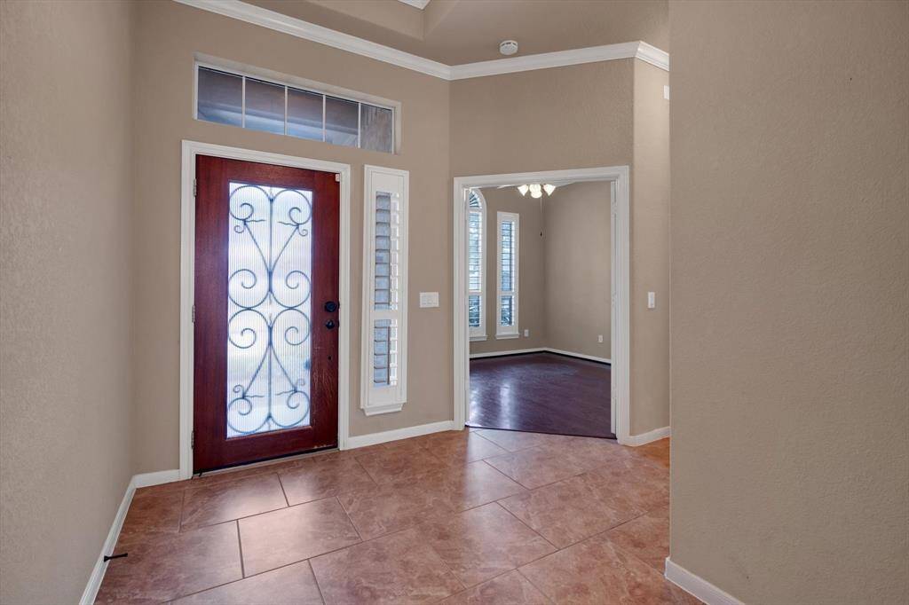 This entryway features a decorative glass front door, tiled flooring, and leads into a room with wood flooring and ample natural light from large windows. The space is accented with crown molding for a touch of elegance.