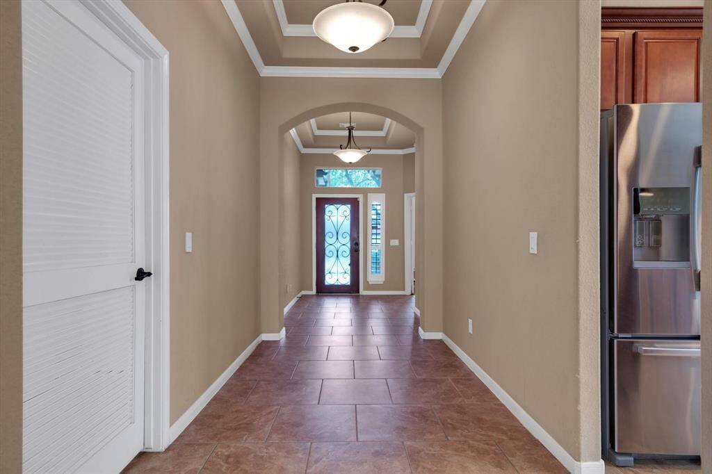 This entryway features a welcoming arched hallway with tile flooring and neutral walls. It leads to a decorative front door with glass and wrought iron accents. The ceiling has recessed lighting and crown molding, adding elegance. A glimpse of the kitchen reveals stainless steel appliances.