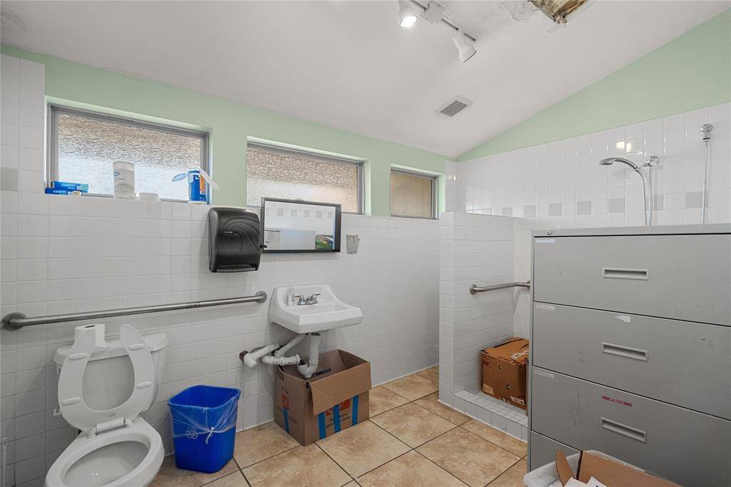 This wheelchair-accessible bathroom oiffers a toilet, sink, and shower. The walls and floor are tiled, and there are handrails for support. The space is light and bright due to the horizontal windows!
