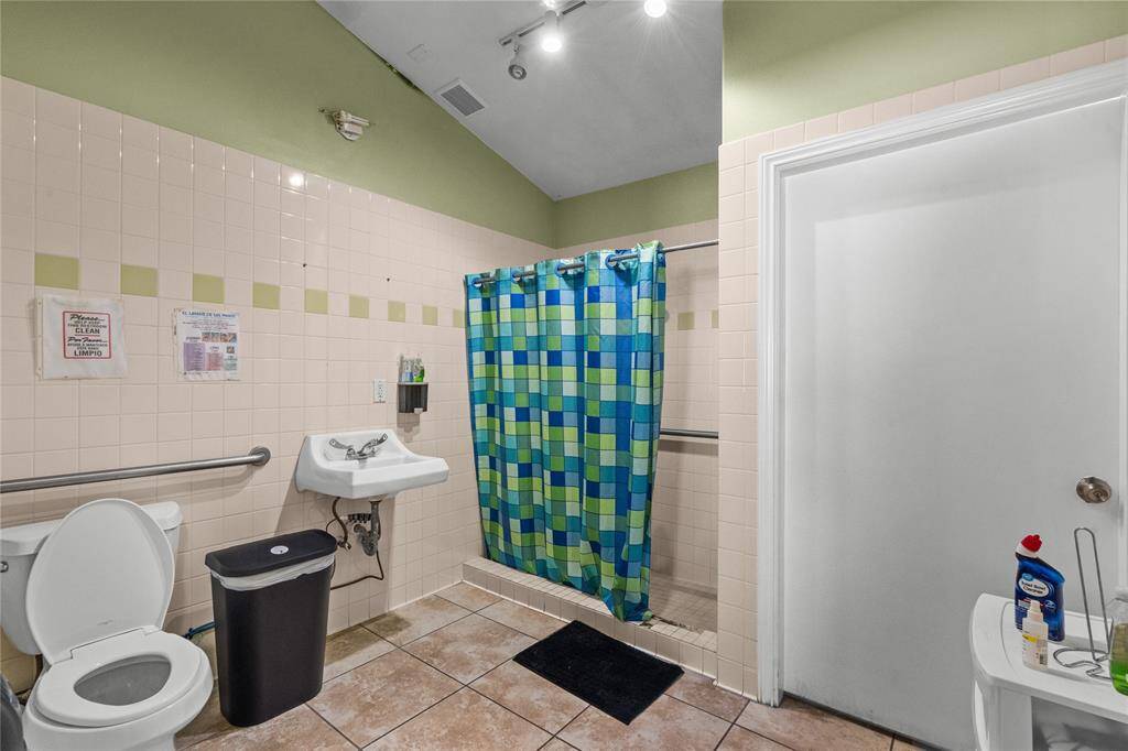 This commercial-grade bathroom features a shower stall, a sink with a soap dispenser, and a toilet. This well maintained space offers tiled walls and floors, and it includes handrails for accessibility.