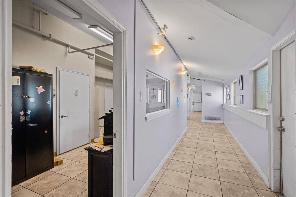 This functional and well-lit space features light walls and a neutral tile floor. There are several doors along one side of the hallway, leading to different rooms. On the opposite side, windows flood the space with natural light.