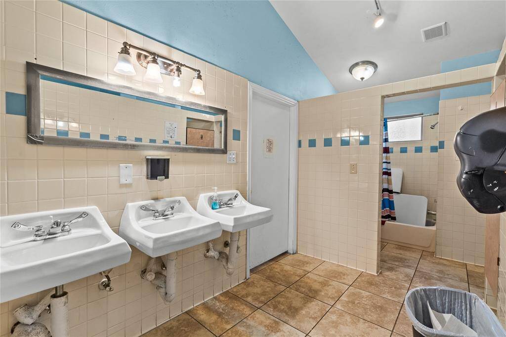 This well-lit bathroom offers three separate sinks with a long mirror above them, an updated lighting fixture, and shower stalls. The walls and floor are tiled in a neutral color, giving the space a clean look.