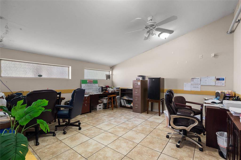 The room off of the entrance features tile flooring, neutral walls, and ample natural light from horizontal windows, making it a practical area for office or workspace setups. Currently arranged as an administrative office with desks, filing cabinets, and storage, this room demonstrates the property’s adaptability.