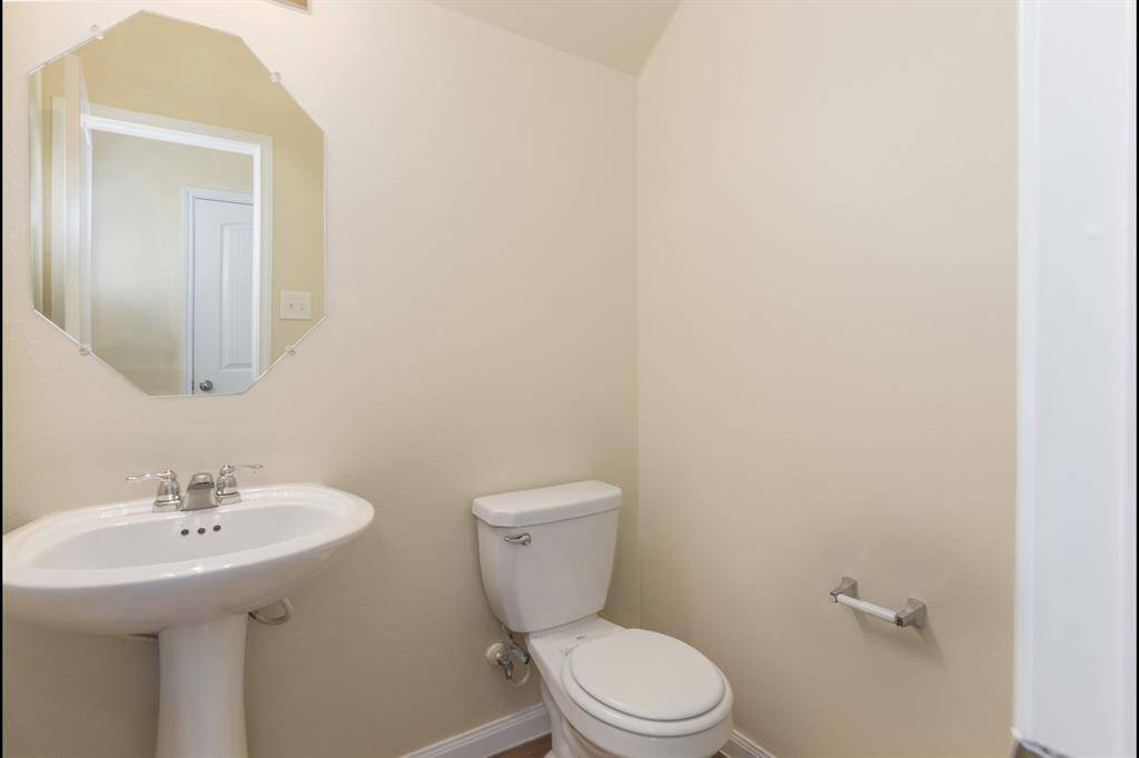 This is a small, well-lit bathroom with a pedestal sink, toilet, and an octagonal mirror. The walls are painted a light, neutral color, and the space appears clean and functional.