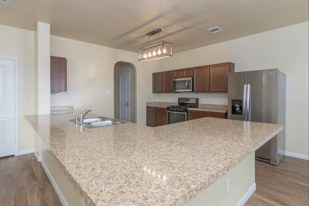 Spacious kitchen with a large, granite-style countertop and modern appliances, including a stainless steel refrigerator and oven. Warm wood cabinetry complements the neutral walls and wooden flooring.