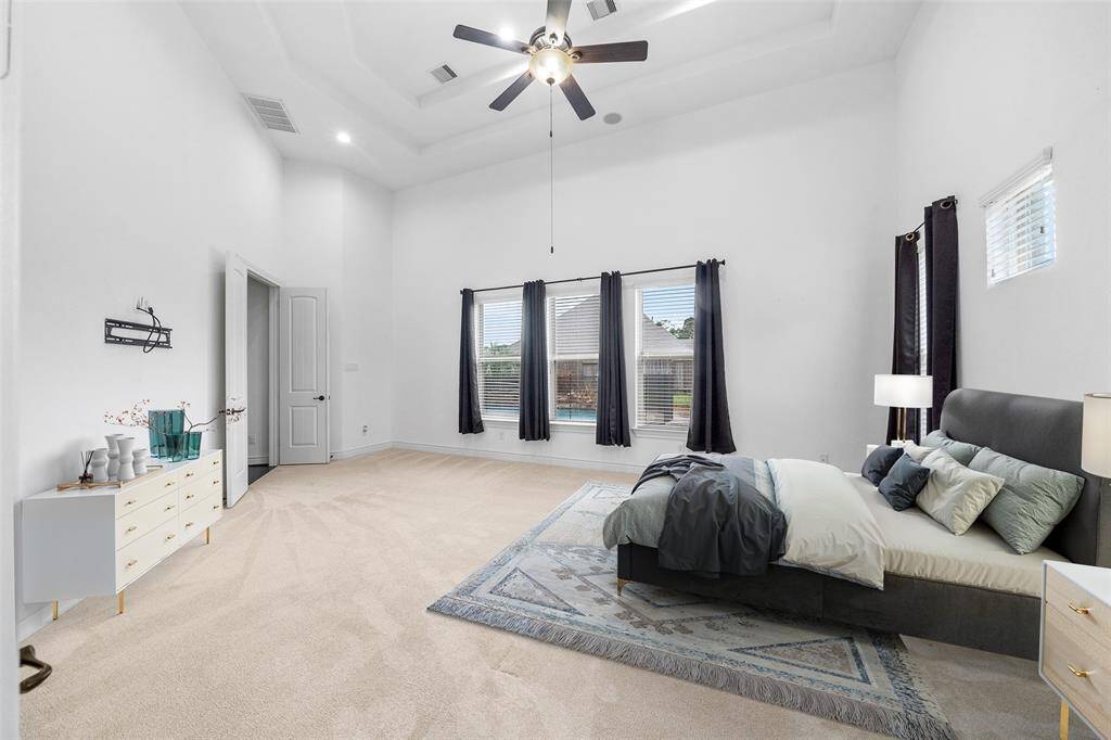 Spacious, bright Primary bedroom with soaring ceilings and neutral tones. Features large windows with dark curtains, a stylish ceiling fan, and plush carpeting. The room is elegant offering a serene and modern retreat. Digitally staged.