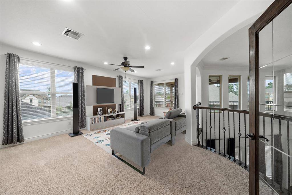 Spacious and bright upstairs living area with large windows, neutral carpeting, a ceiling fan, and a cozy seating arrangement. The room features a modern entertainment setup and overlooks a scenic neighborhood view. The open layout is complemented by elegant railings and an arched doorway. This game room has access to the large outdoor balcony. Digitally staged