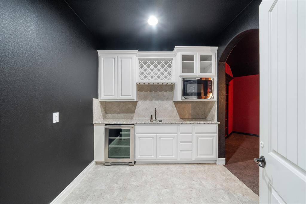 The upstairs refreshment/  wet bar area with sleek white cabinetry, a built-in microwave, and a wine rack. The granite countertop includes a small sink, and there's a mini-fridge below. The space features dark walls and bright flooring, creating a modern contrast.