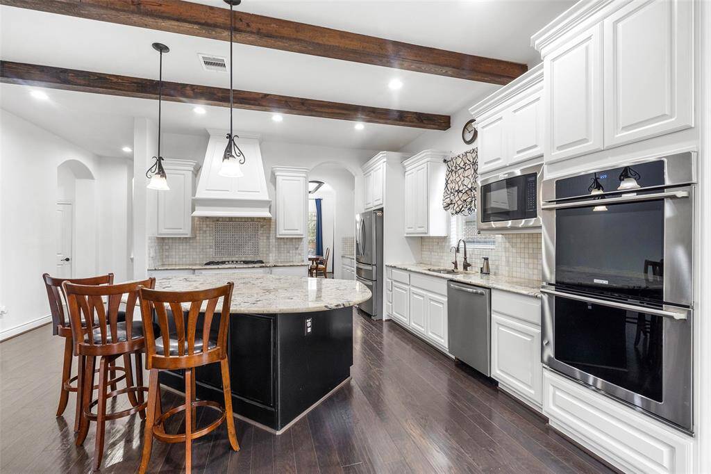The chef's kitchen features elegant dark wood floors with exposed ceiling beams, white custom cabinetry, granite countertops, and a huge central island with seating. The modern stainless steel appliances, including double ovens, and stylish light fixtures complete the look.