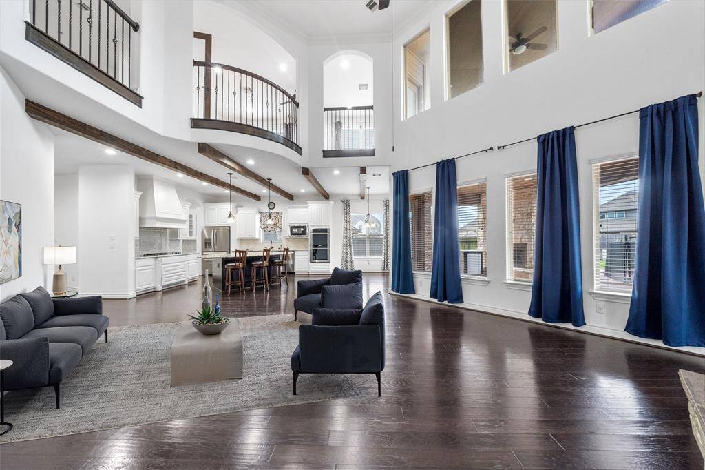 The spacious living area features soaring ceilings and large windows that fill the room with natural light. It is open to the kitchen and breakfast area. The upper-level balcony adds architectural interest and overlooks the expansive living space. Digitally staged