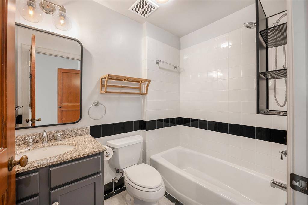 This is a clean and well-maintained bathroom featuring a shower/tub combo with black and white tiling, a single vanity with granite countertop, and modern lighting fixtures above a large mirror.
