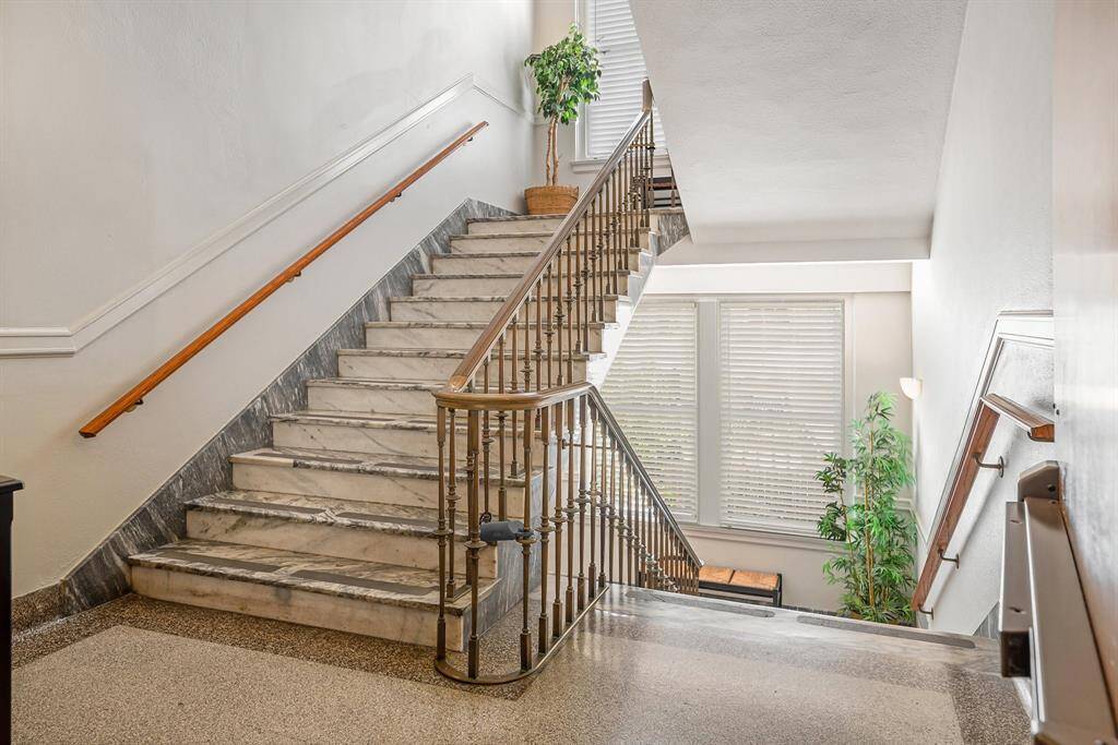 The photo showcases the elegant interior common staircase with marble steps, complemented by a classic metal railing and brass handrail. The space is well-lit with natural light filtering through the window, and there's a touch of greenery from a potted plant enhancing the welcoming ambiance.
