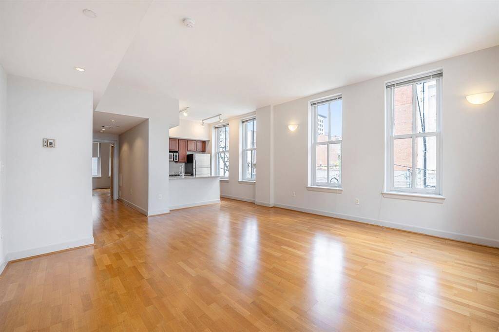 This is a bright, open-concept living space featuring hardwood floors, multiple large windows with abundant natural light, and an adjacent kitchen with modern cabinetry. The area is spacious and has high ceilings, ideal for both living and entertaining.