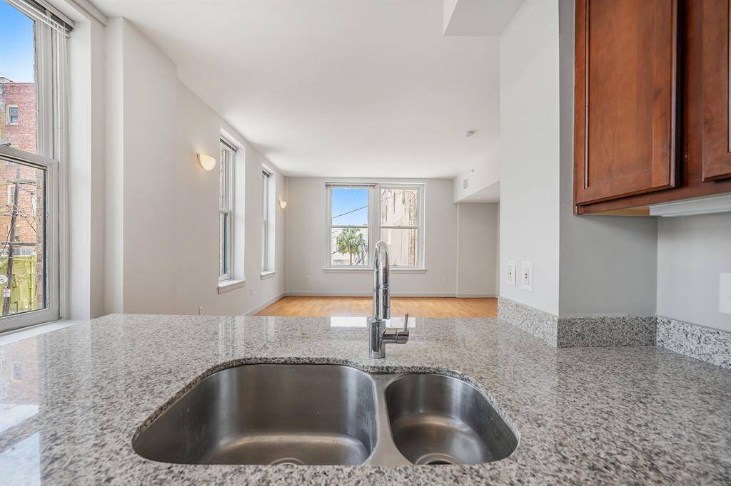 This photo showcases a bright, modern kitchen with stainless steel double sink, granite countertops, wood tone cabinetry, and hardwood floors.