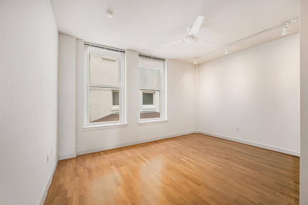 This is the primary bedroom with hardwood floors, two large windows for natural light, a ceiling fan, and track lighting. The walls are painted light gray, creating a clean, blank canvas for personalization.