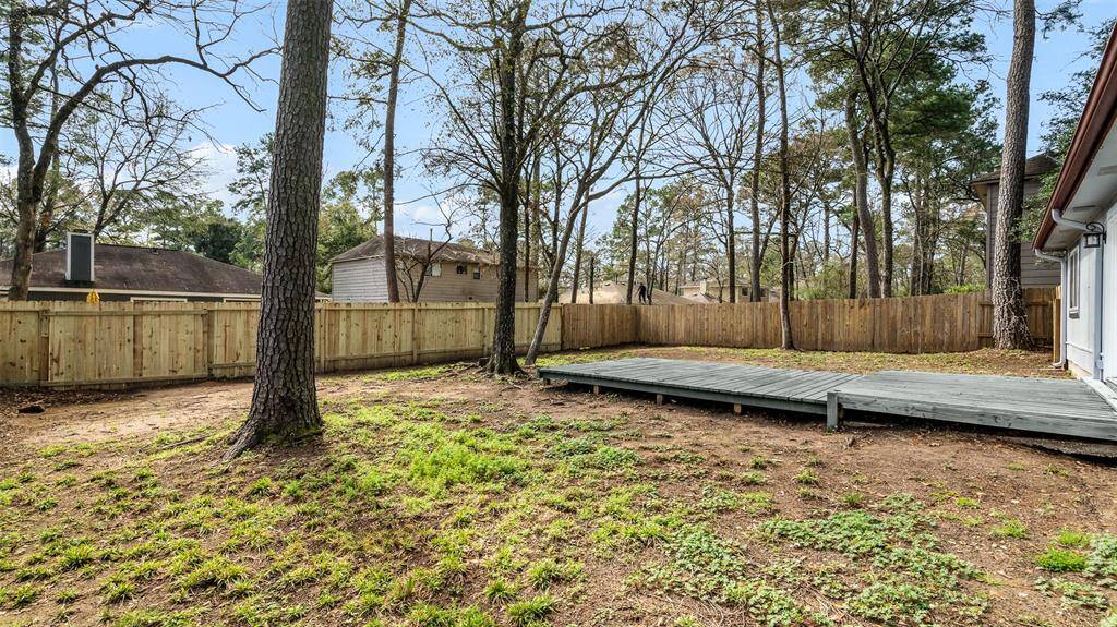 Backyard with wooden deck, surrounded by a wooden fence. The yard has sparse grass and a few tall trees, providing a natural setting. It's adjacent to a house with light-colored siding and features an open, spacious feel.