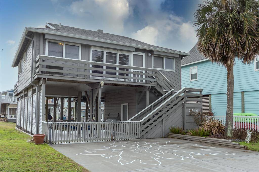 This is a two-story coastal canal home with a large balcony and an hardi plank exterior. It features an elevated main living area accessible by stairs, and a covered space underneath, ideal for parking or outdoor activities. The property is accented by a palm tree and a corner lot