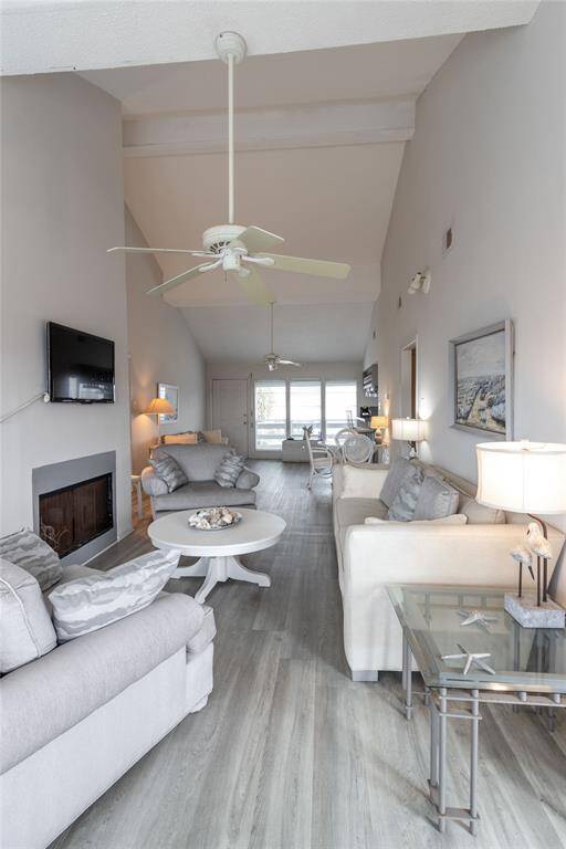 This bright, open-concept living room features vaulted ceilings, neutral tones, and vinyl plank flooring. It includes comfortable seating, a mounted TV, fireplace (not working, and large windows that let in plenty of natural light. The space is stylishly decorated, promoting a cozy and inviting atmosphere.