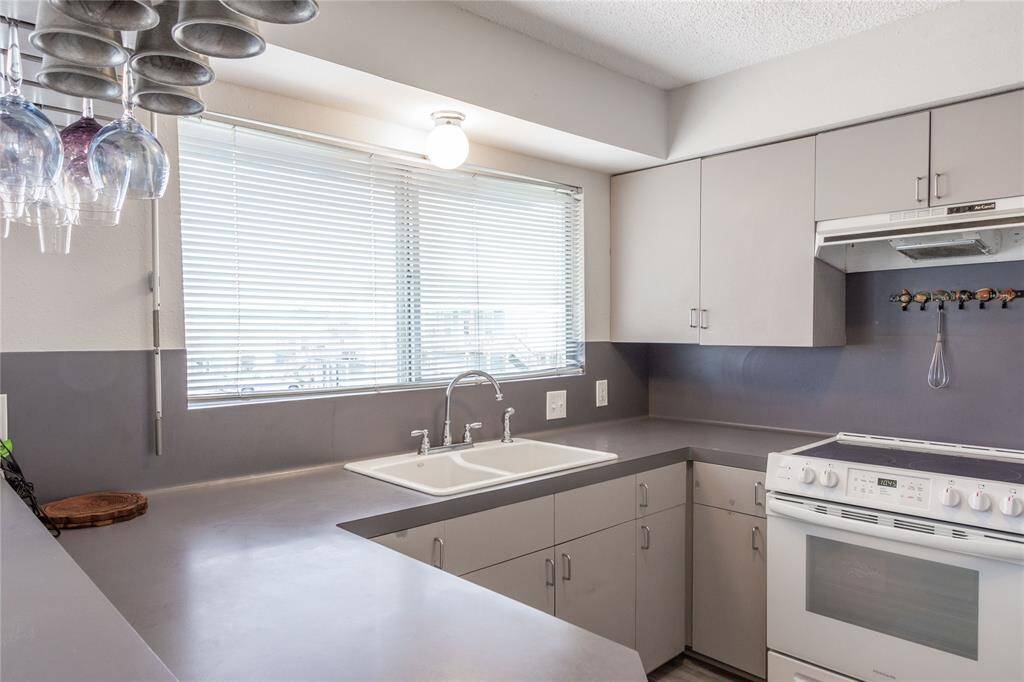 Modern kitchen with sleek gray countertops and cabinets, featuring a double sink under a large window. There's a white oven and overhead storage, with hanging wine glasses adding a decorative touch. Bright and functional space. New kitchen sink and faucet