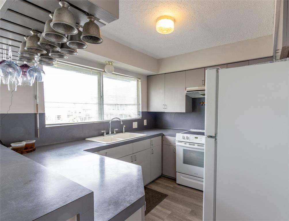 This kitchen features a modern design with ample natural light, thanks to large windows. It includes a white stove and refrigerator, grey countertops, and plenty of cabinet space. The overhead glass and mug rack adds a unique touch. Pewter items do not remain