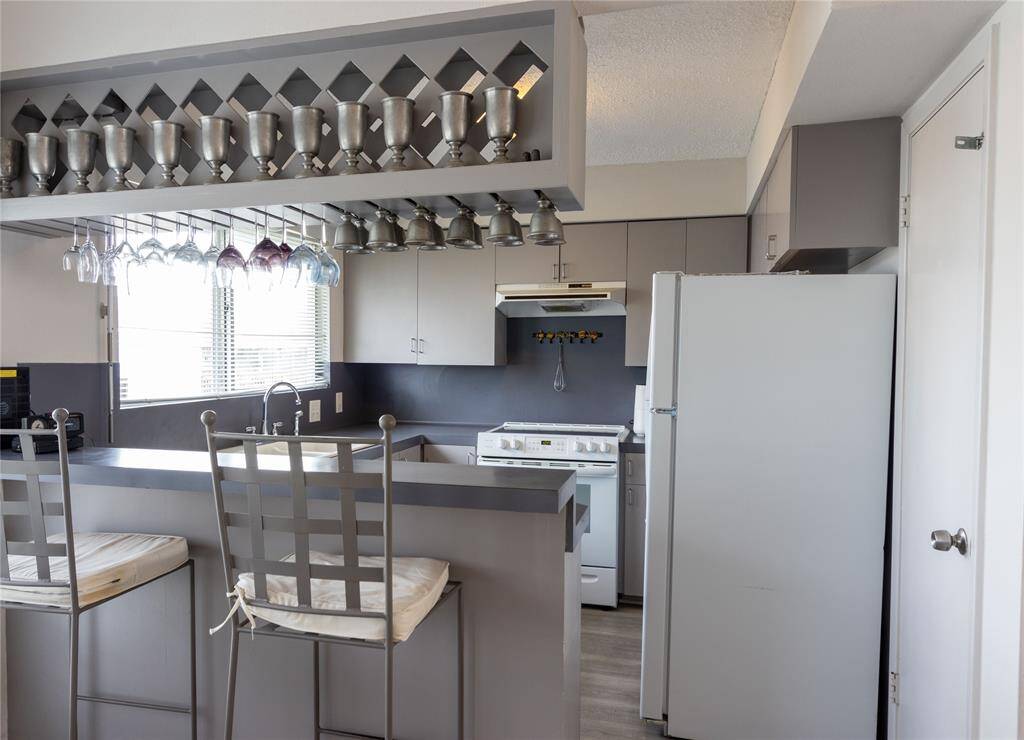 Modern kitchen with sleek gray cabinetry and countertops. Features a breakfast bar with seating for two and overhead glass storage. Includes a large window for natural light and essential appliances. Pewter items do not remain.