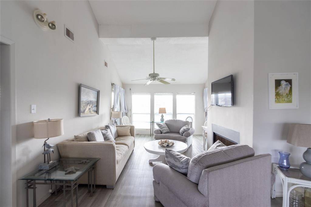 Bright and airy living room with high ceilings and neutral tones. Features a cozy seating area, large windows allowing natural light, a ceiling fan, and a wall-mounted TV. Ideal for relaxation and entertaining.