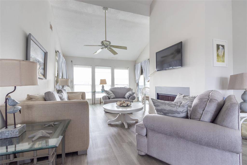 Bright and airy living room with high ceilings, large windows, and neutral decor. Features include comfortable seating, a wall-mounted TV, and a ceiling fan. A fireplace that seller does not use. Ideal for relaxation and gatherings.