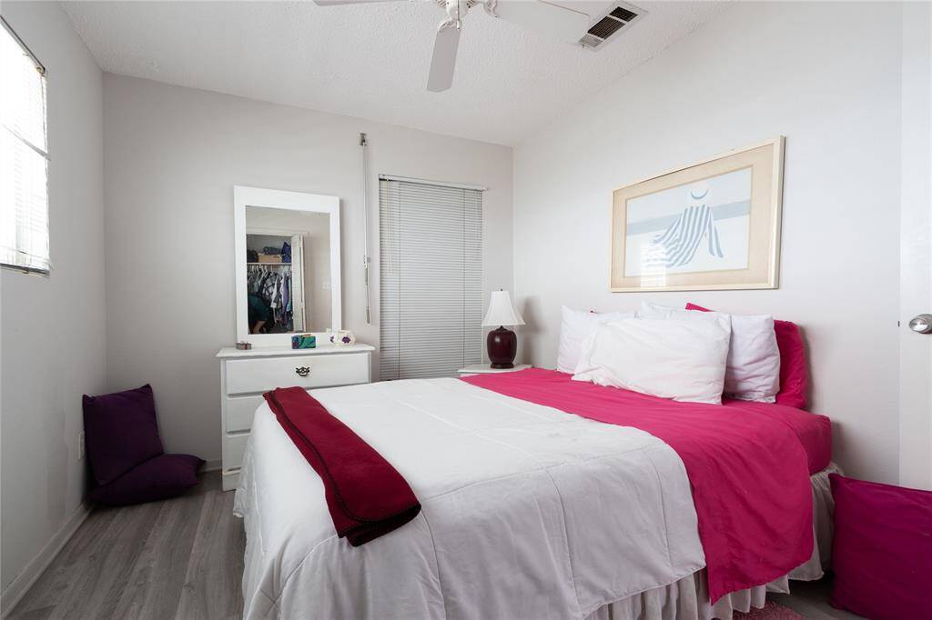 Cozy bedroom featuring a comfortable bed with a white dresser with a mirror, and a small bedside lamp. The room has a modern feel with neutral walls, and natural light from a window with blinds.