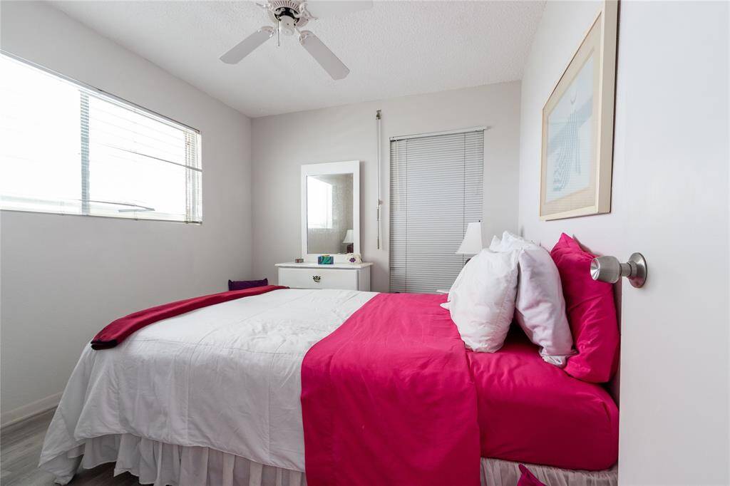 This cozy bedroom features a bright and inviting atmosphere with a large window, ceiling fan, and vibrant  bedding. It includes a small dresser with a mirror and a framed artwork on the wall, creating a comfortable and stylish space.