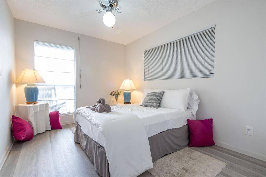 This cozy bedroom features a comfortable bed with white and gray linens, accented by vibrant pillows. Natural light streams through a large window, complemented by stylish lamps on each side. The room has a light, neutral color palette and modern vinyl plank flooring, offering a welcoming and serene atmosphere.