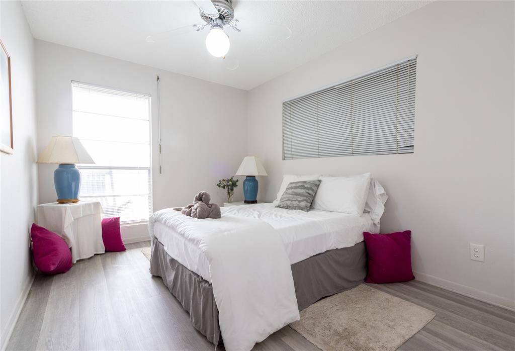 Cozy bedroom with a neutral color palette, featuring a comfortable bed, modern ceiling fan, and large windows for natural light. Accents include bright pillows and blue lamps, creating a welcoming atmosphere.