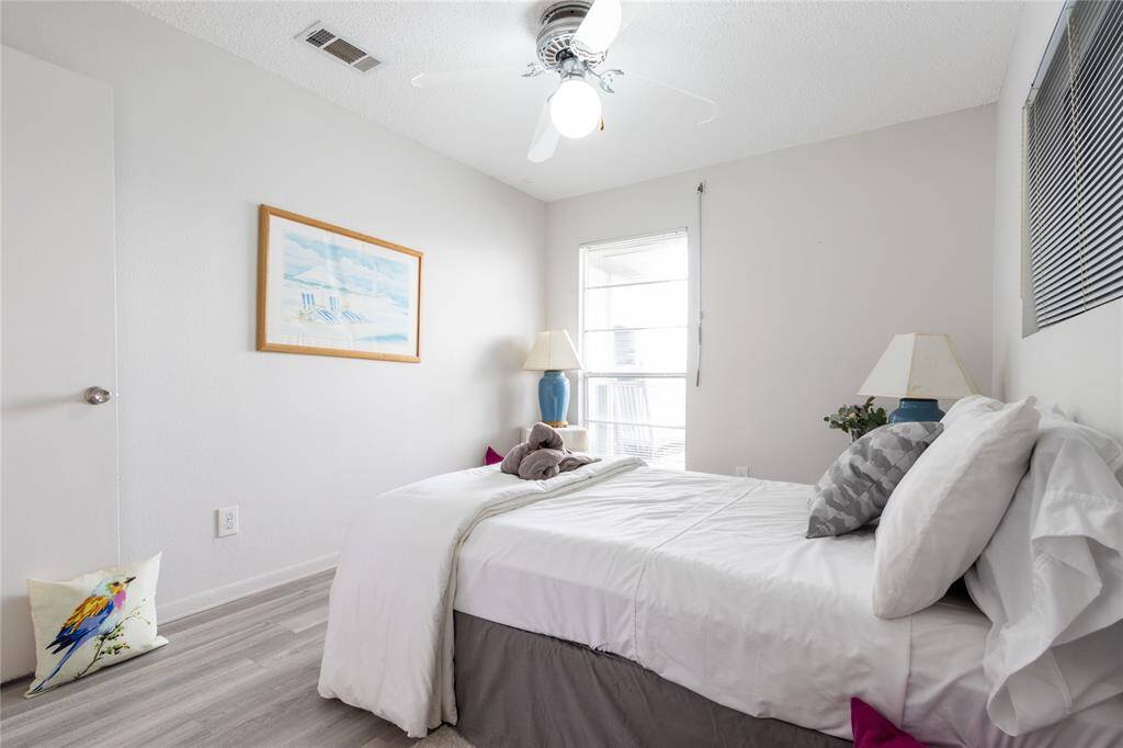 This is a bright, cozy bedroom featuring light-colored walls and vinyl plank flooring. It includes a bed with neutral bedding, a ceiling fan, and a large window that allows natural light. The room is simply decorated with a framed picture, a bedside lamp, and a decorative pillow.
