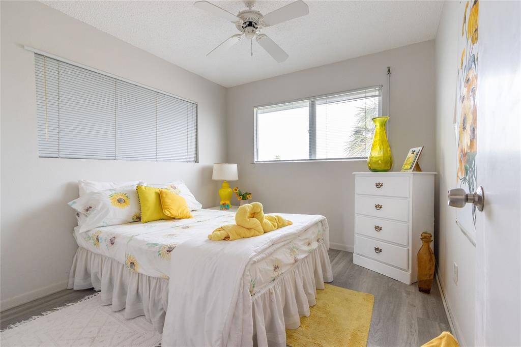 Cozy bedroom with bright natural light, featuring a comfortable bed with floral bedding, a ceiling fan, and a white dresser. The room is accented with cheerful yellow decor, creating a warm and inviting atmosphere. Sunflower painting does not remain.