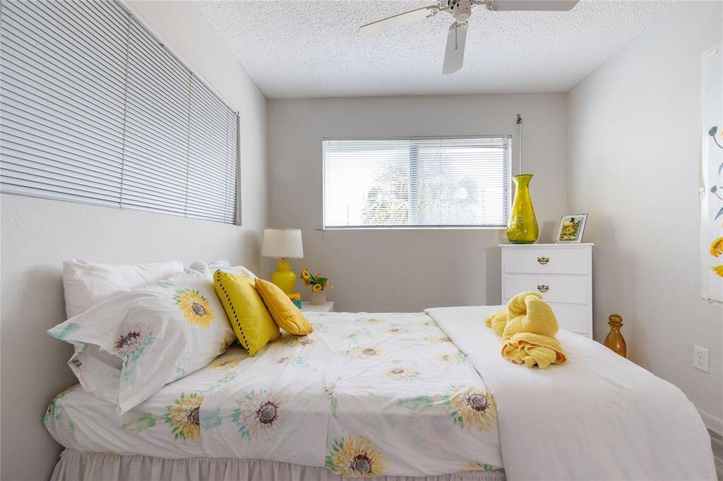 A cozy bedroom with a floral-themed bed, neutral walls, and two large windows for natural light. It features a ceiling fan, a white dresser with decorative accents, and cheerful yellow tones throughout.