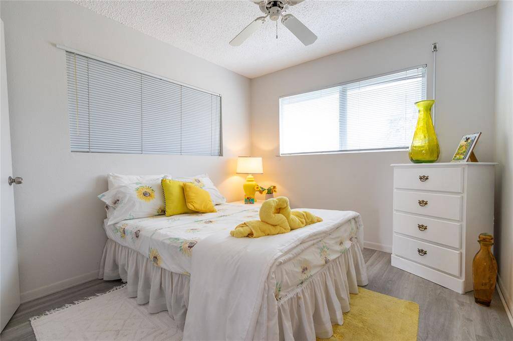 This cozy bedroom features a soft, neutral color palette with a comfortable bed, floral bedding, and bright yellow accents. It includes a ceiling fan, ample natural light from two windows, and a white dresser for storage. The space is inviting and well-lit, perfect for relaxation. Vinyl plank flooring.