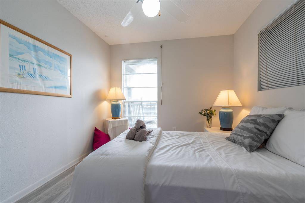 This cozy bedroom features a comfortable bed with neutral bedding, two bedside tables with lamps, a large window letting in natural light, and a serene beach-themed artwork on the wall.