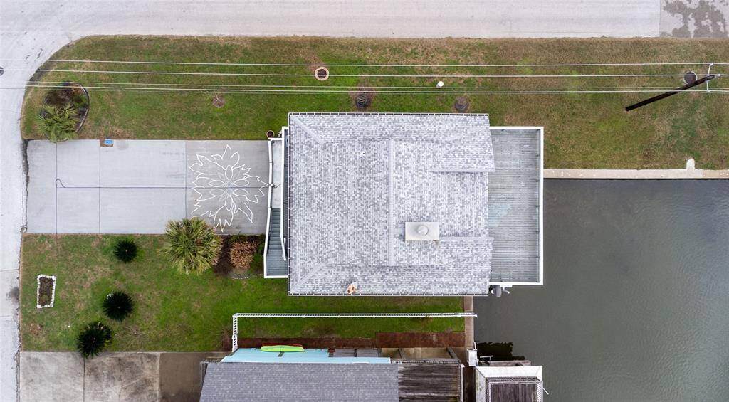 This aerial photo shows a waterfront home with a gray roof, adjacent to a canal. The property features a paved driveway with decorative hand painted art and a small, landscaped front yard. Corner lot