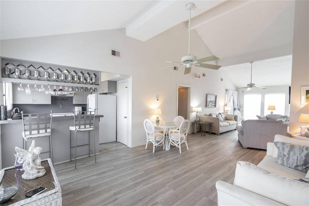 This spacious living area features high ceilings and an open layout, seamlessly connecting the kitchen and living room. The kitchen includes a small bar area with seating, and the space is filled with natural light from large windows. Neutral tones and modern finishes create a warm, inviting atmosphere.