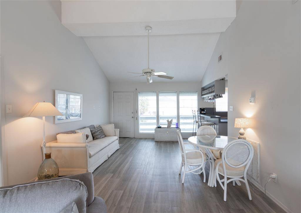 This bright, open-concept living space features high ceilings, light vinyl plank flooring, and ample natural light from large windows. It includes a cozy seating area, dining set, and a modern kitchen with a convenient breakfast bar.