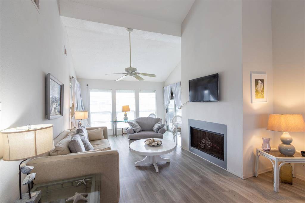 Bright and airy living room with high ceilings, featuring a cozy fireplace (not working), ceiling fan, and ample natural light from large windows. The space is tastefully furnished with neutral tones, creating a welcoming atmosphere.
