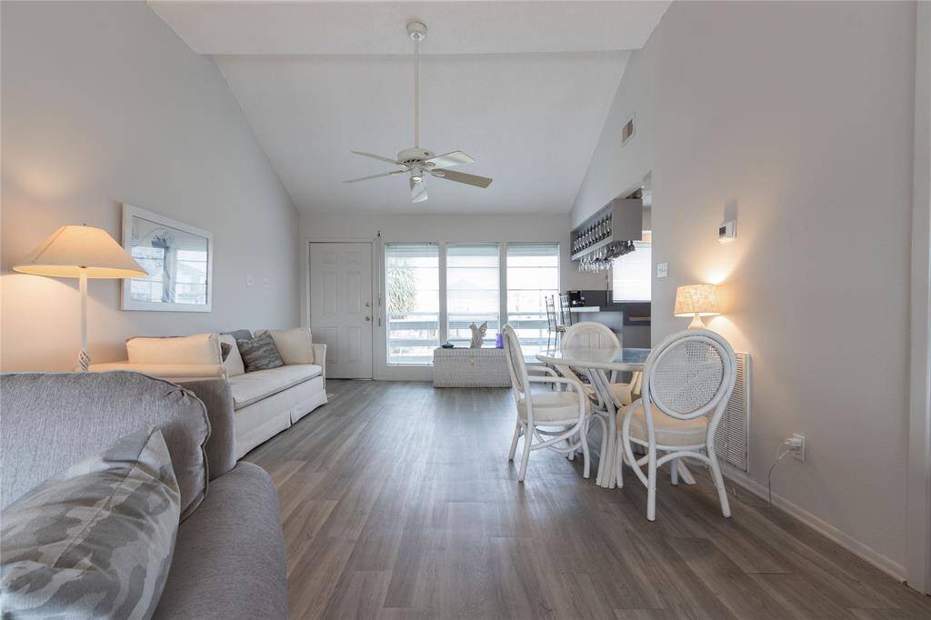 This bright living space features high ceilings with a fan, light vinyl plank flooring, and a neutral color palette. It includes a cozy seating area, a small dining table, and large windows providing ample natural light.