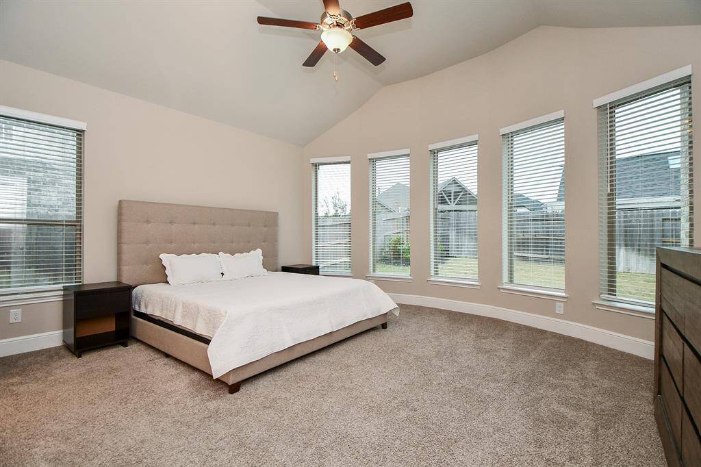 Primary suite  features a high ceiling with a fan, large windows allowing ample natural light, neutral walls, and plush carpeting, creating a cozy and inviting atmosphere.