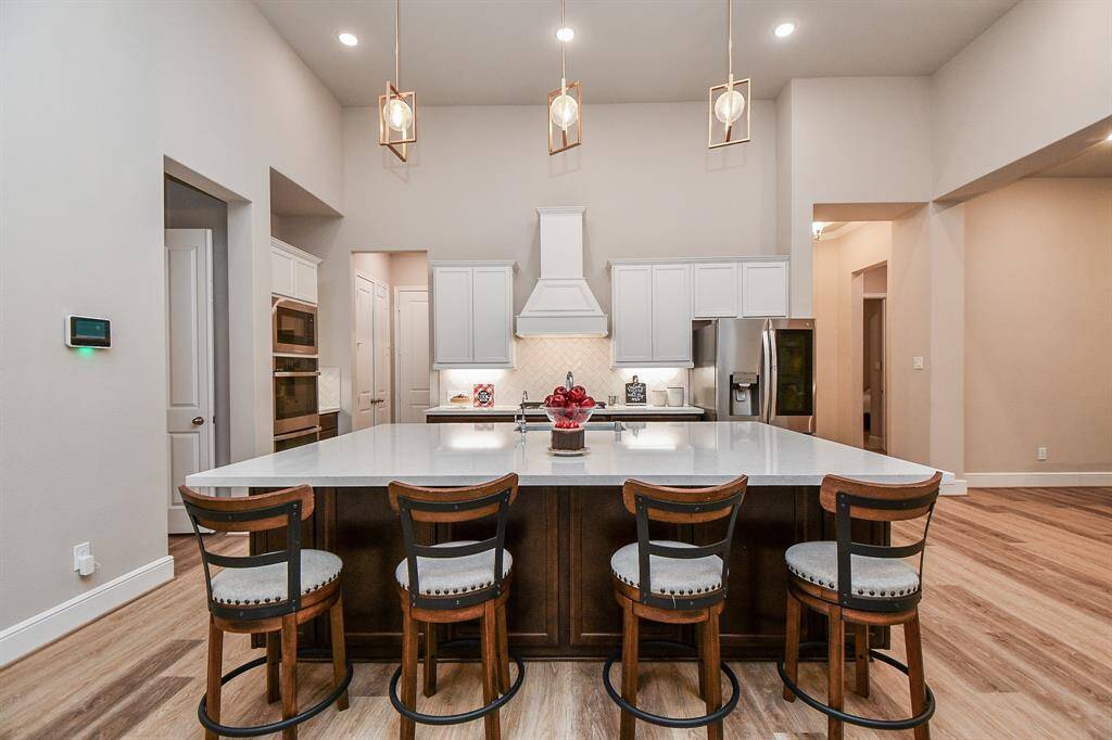 This kitchen features an expansive island with seating for four, modern pendant lighting, stainless steel appliances, and sleek white and wood stained cabinetry. Its open layout and warm luxury vinyl flooring create a welcoming atmosphere.
