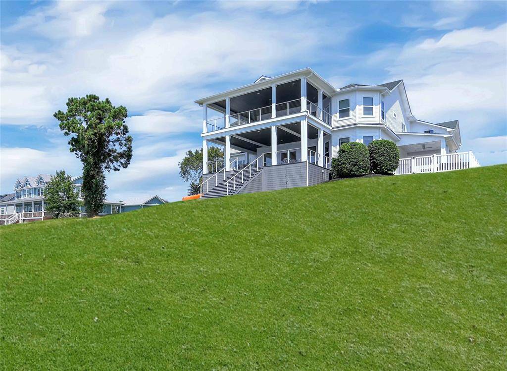 This photo showcases a spacious, modern home with a large, elevated deck and multiple stories. It sits on a gentle hill with expansive grassy lawn, offering picturesque views and ample outdoor space. The house features large windows and a wraparound porch, adding to its charm and appeal.
