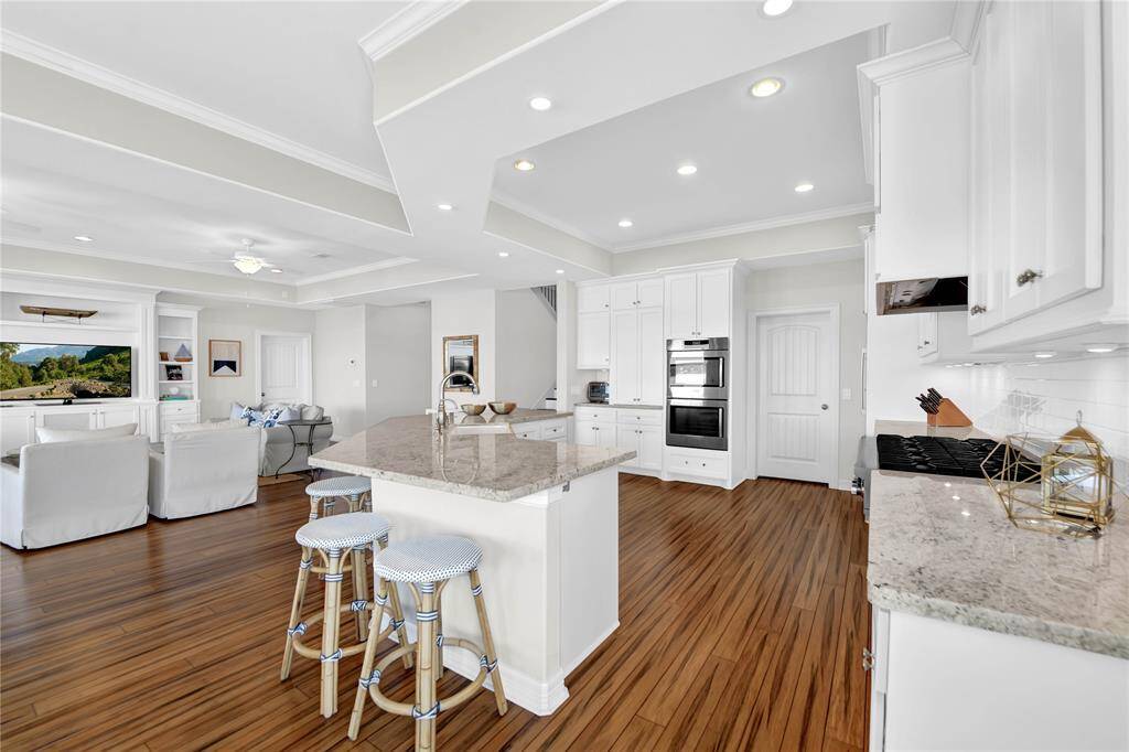 This photo showcases a spacious, open-concept kitchen and living area with hardwood floors. The kitchen features white cabinetry, granite countertops, and modern appliances, including a double oven. An island with seating enhances the space, which flows into a comfortable living area with built-in shelving and a large window offering scenic views.