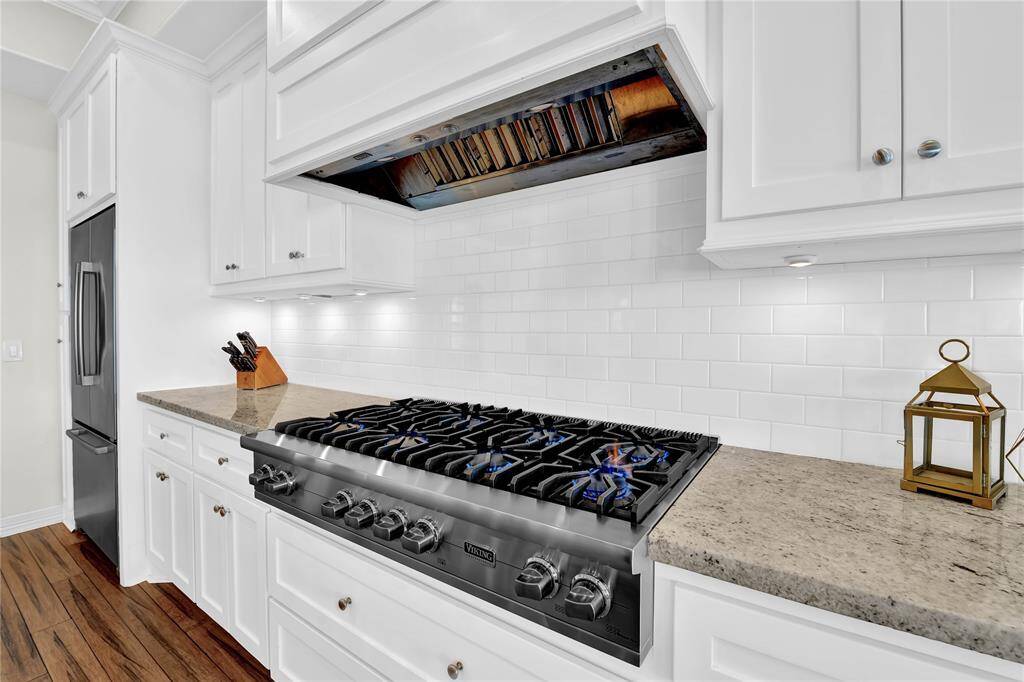 This kitchen features sleek white cabinetry, granite countertops, and a high-end gas stove with a modern hood. The stainless steel refrigerator and subway tile backsplash add a contemporary touch, complemented by wood flooring.