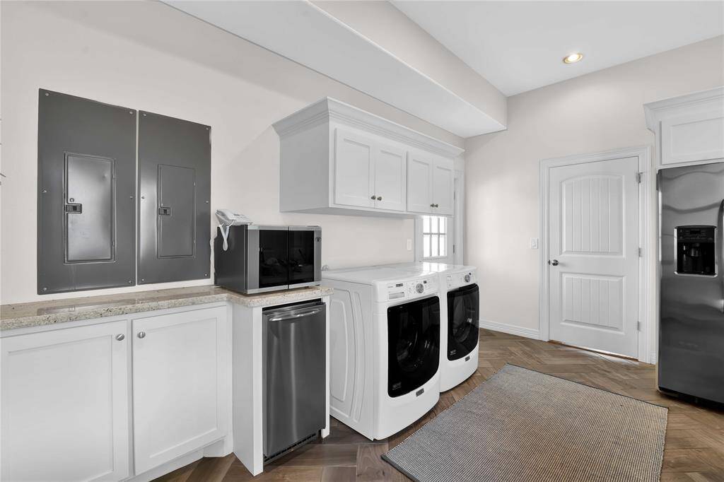This photo showcases a modern laundry room with white cabinetry, a washer and dryer, a countertop with a microwave, and a sleek stainless steel fridge. The room features ample storage, neutral tones, and a herringbone-patterned floor.