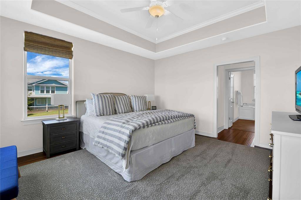 This photo shows a spacious bedroom with a large bed, tray ceiling, and neutral walls. It features a window with natural light, a ceiling fan, and an adjacent bathroom. The room has a cozy carpet and a modern feel, perfect for relaxation.